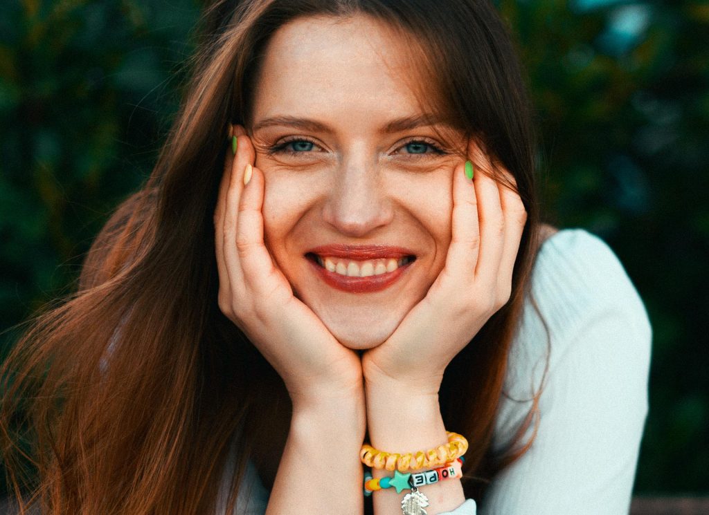 Lächelnde Frau mit langen, braunen Haaren, die ihre Hände an das Gesicht stützt. Sie trägt ein helles Oberteil und bunte Armbänder. Der Hintergrund zeigt grüne Pflanzen und vermittelt eine fröhliche, entspannte Stimmung.