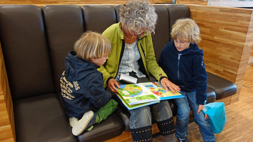 Eine ältere Frau sitzt auf einer Bank und liest mit zwei Kindern ein buntes Bilderbuch. Die Kinder sind aufmerksam und zeigen Interesse an den Illustrationen. Im Hintergrund ist eine moderne Umgebung mit Holzdekor zu sehen.