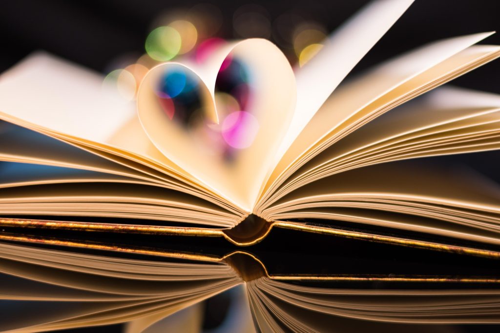 Offenes Buch mit einer Seitenform, die ein Herz bildet, vor einem verschwommenen, bunten Hintergrund. Ideal für Themen wie Liebe, Bücher und Kreativität.