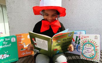 Ein Kind im Kostüm des Katers mit Hut liest ein Buch von Dr. Seuss. Um es herum sind weitere Bücher von Dr. Seuss und P.D. Eastman aufgestellt. Die bunte Umgebung und das fröhliche Outfit fördern die Lesefreude und Kreativität.