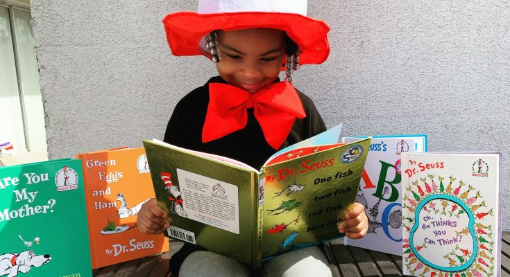 Ein Kind im Kostüm des Katers mit Hut liest ein Buch von Dr. Seuss. Um es herum sind weitere Bücher von Dr. Seuss und P.D. Eastman aufgestellt. Die bunte Umgebung und das fröhliche Outfit fördern die Lesefreude und Kreativität.