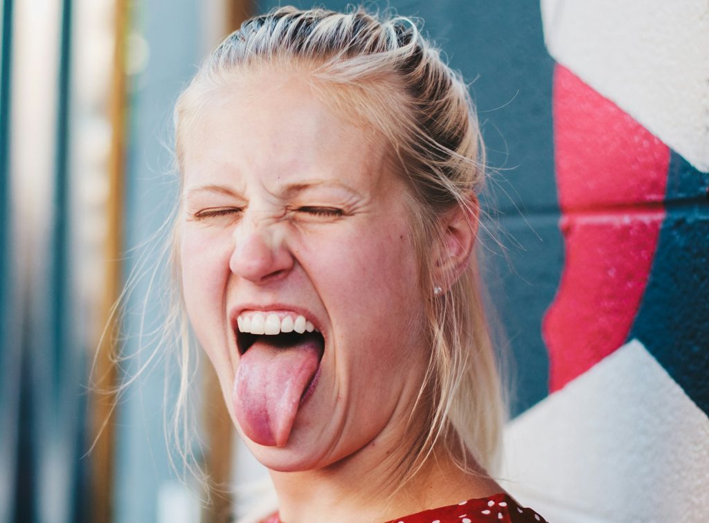 Junge Frau mit blonden Haaren, die die Augen zusammenkneift und die Zunge herausstreckt, vor einer bunten Wand. Sie trägt ein rotes Kleid mit weißen Punkten und zeigt eine fröhliche, verspielte Ausdrucksweise.