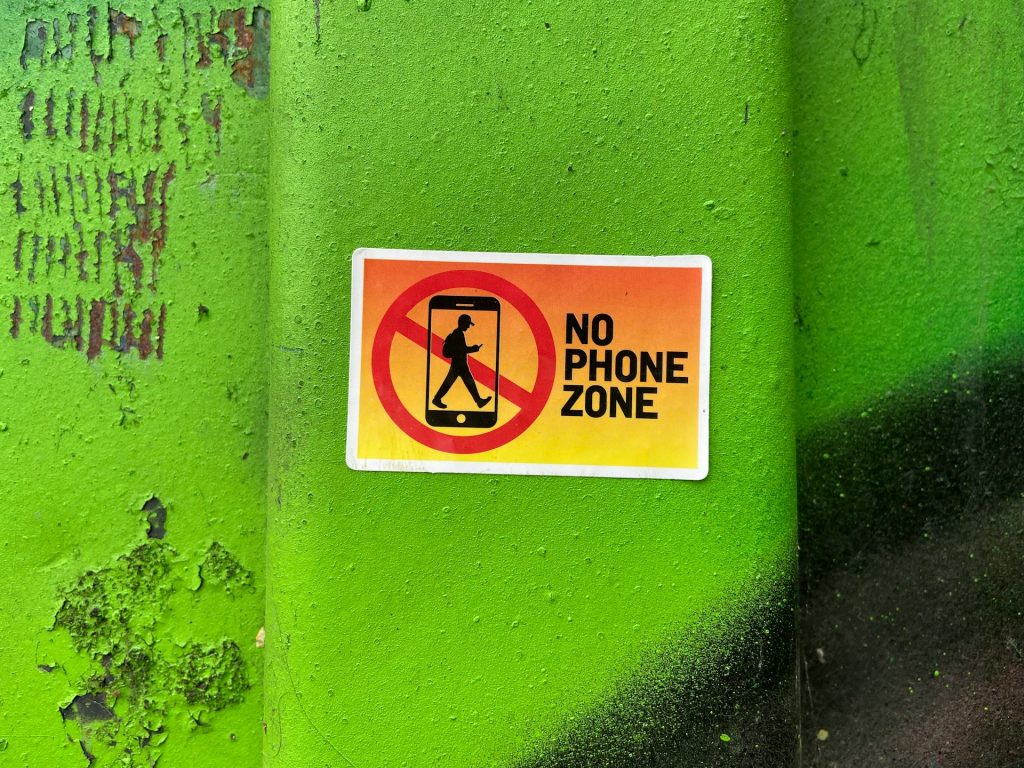Auf einem grellgrünen Hintergrund ist ein Aufkleber mit der Aufschrift "No Phone Zone" zu sehen, der darauf hinweist, dass Handynutzung in diesem Bereich nicht erlaubt ist. Der Aufkleber zeigt eine durchgestrichene Smartphone-Silhouette und eine Person, die mit dem Telefon geht.