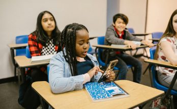 Schülerinnen und Schüler sitzen in einem Klassenzimmer. Eine Schülerin mit geflochtenen Haaren nutzt ihr Smartphone, während die anderen aufmerksam im Hintergrund sitzen. Die Tische sind mit Schulmaterialien ausgestattet, und die Atmosphäre wirkt konzentriert.