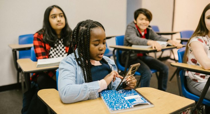 Schülerinnen und Schüler sitzen in einem Klassenzimmer. Eine Schülerin mit geflochtenen Haaren nutzt ihr Smartphone, während die anderen aufmerksam im Hintergrund sitzen. Die Tische sind mit Schulmaterialien ausgestattet, und die Atmosphäre wirkt konzentriert.