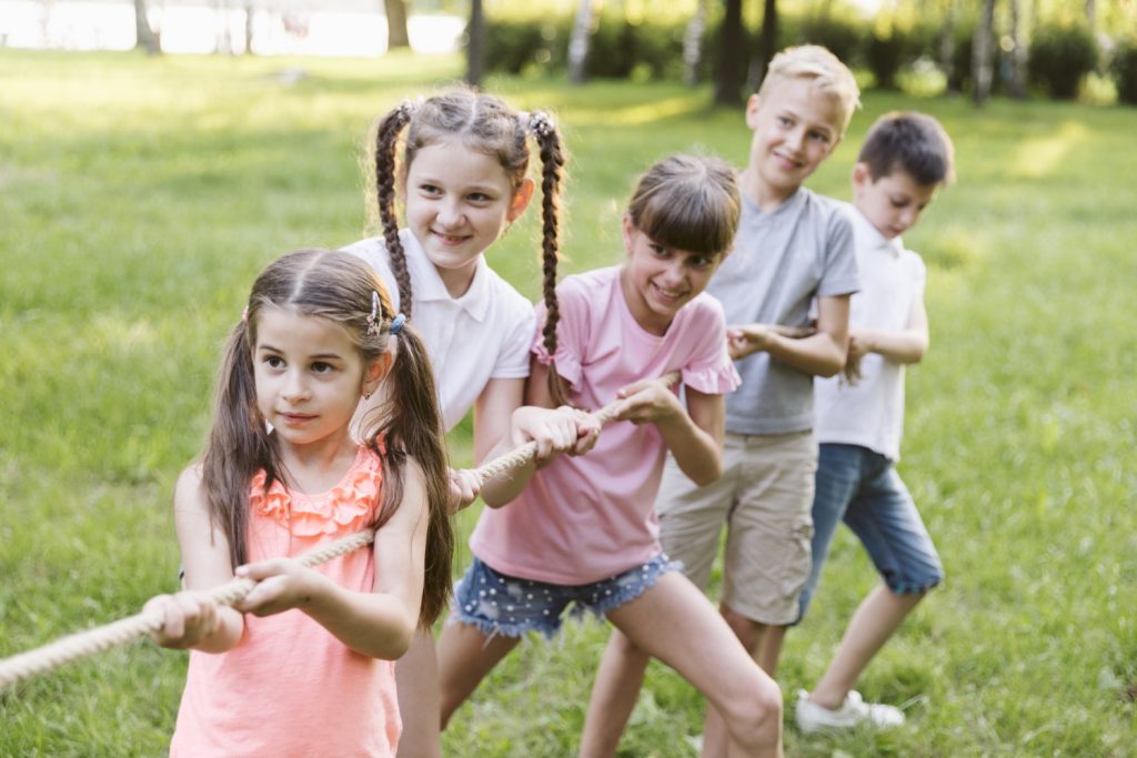 Fünf Kinder ziehen gemeinsam an einem Seil im Freien, umgeben von einer grünen Wiese. Sie zeigen Freude und Teamarbeit während eines Spiels oder Wettbewerbs.