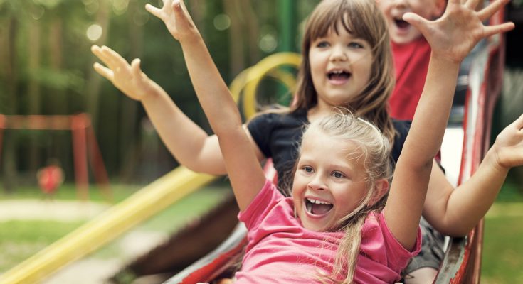 Kinder haben Spaß auf einer Rutsche im Spielplatz, während sie fröhlich ihre Hände in die Luft werfen. Der Hintergrund zeigt eine grüne, natürliche Umgebung mit Spielgeräten.