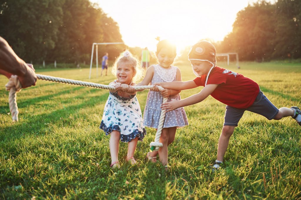 Kinder spielen im Freien ein Seilziehen-Spiel auf einer Wiese, umgeben von Sonnenlicht. Drei fröhliche Kinder, zwei Mädchen und ein Junge, ziehen gemeinsam an einem dicken Seil. Im Hintergrund sind weitere Kinder beim Fußballspielen zu sehen.