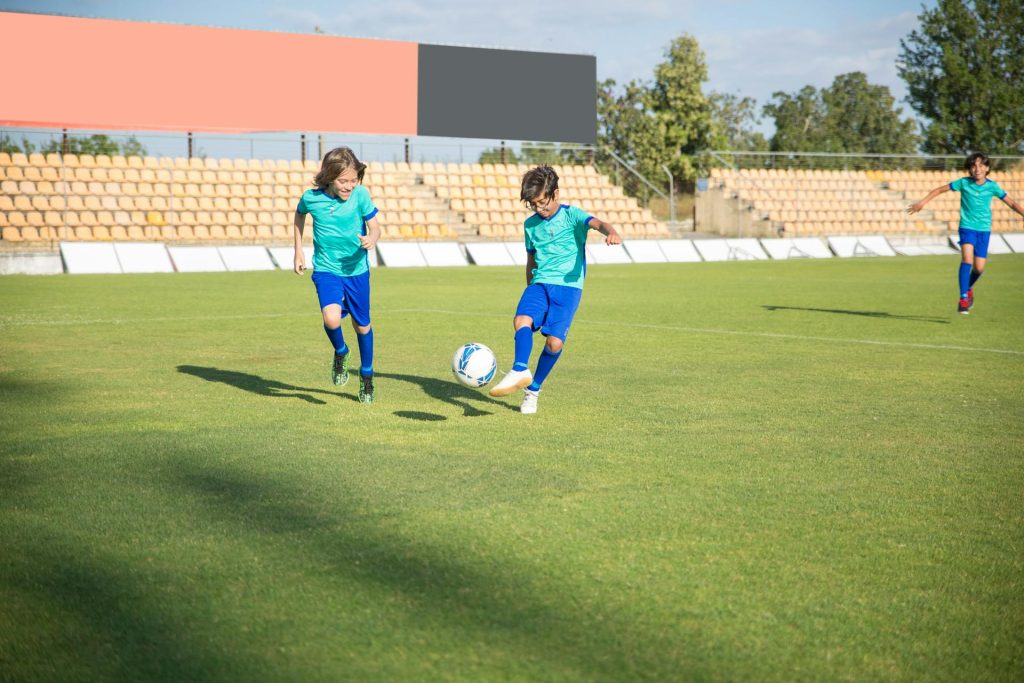 Zwei Kinder in blauen Fußballtrikots spielen auf einem grünen Fußballfeld. Einer der Jungen dribbelt den Ball, während der andere ihm mit Freude nachläuft. Im Hintergrund sind leere Stadionplätze sichtbar.