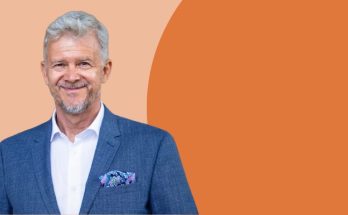 Ein älterer Mann mit grauen Haaren trägt einen blauen Anzug und ein weißes Hemd. Er lächelt freundlich in die Kamera und hat ein buntes Einstecktuch. Im Hintergrund sind sanfte, warme Farben zu sehen, die eine einladende Atmosphäre schaffen.