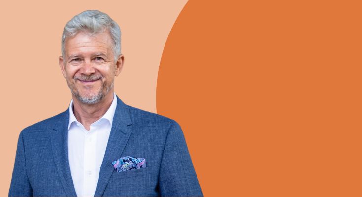 Ein älterer Mann mit grauen Haaren trägt einen blauen Anzug und ein weißes Hemd. Er lächelt freundlich in die Kamera und hat ein buntes Einstecktuch. Im Hintergrund sind sanfte, warme Farben zu sehen, die eine einladende Atmosphäre schaffen.