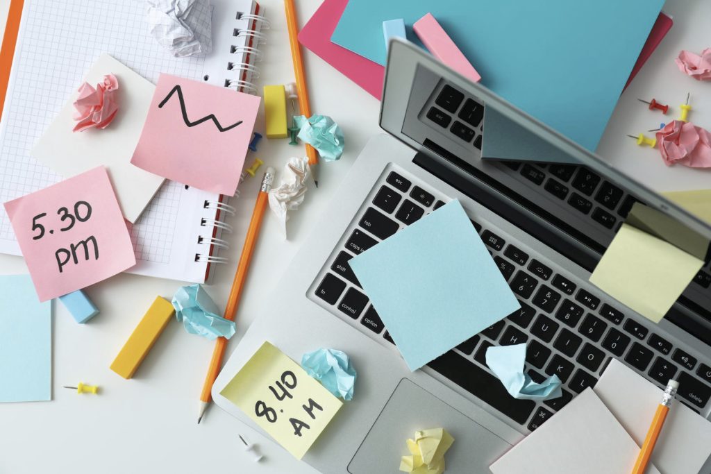 Ein unordentlicher Schreibtisch mit einem Laptop, bunten Haftnotizen, einem Notizbuch und zerknülltem Papier. Auf den Haftnotizen stehen verschiedene Zeiten und Skizzen. Der Tisch ist mit Bürobedarf wie Stiften und Büroklammern bedeckt, was eine kreative Arbeitsumgebung widerspiegelt.