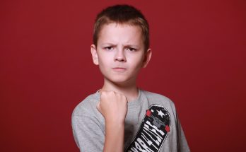 Ein Junge mit kurzem Haar und einem grauen T-Shirt mit Skateboard-Motiv steht vor einem roten Hintergrund. Er zeigt eine entschlossene Pose mit geballter Faust und ernstem Gesichtsausdruck.