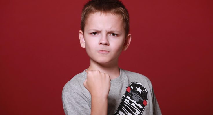 Ein Junge mit kurzem Haar und einem grauen T-Shirt mit Skateboard-Motiv steht vor einem roten Hintergrund. Er zeigt eine entschlossene Pose mit geballter Faust und ernstem Gesichtsausdruck.