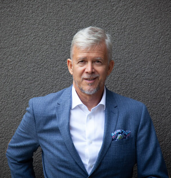 Mann mittleren Alters mit grauen Haaren, der in einem blauen Anzug mit weißem Hemd und farbigem Einstecktuch vor einer grauen Wand steht und freundlich lächelt.