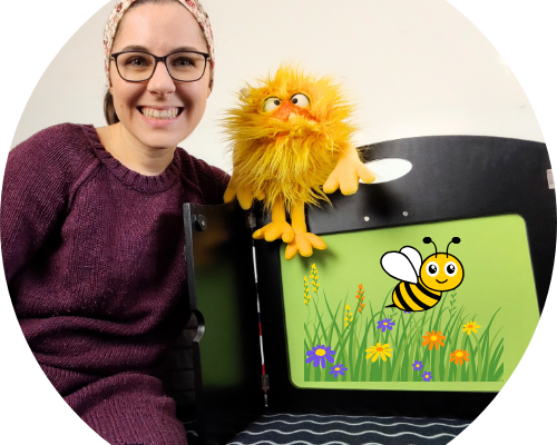 Eine lächelnde Frau mit Brille sitzt neben einer Handpuppe in Form eines gelben Wesens. Im Hintergrund ist eine grüne Tafel mit einer bunten Zeichnung einer Biene und Blumen zu sehen. Die Szene vermittelt eine freundliche und kreative Atmosphäre, ideal für Kinderunterhaltung oder Bildungsinhalte.
