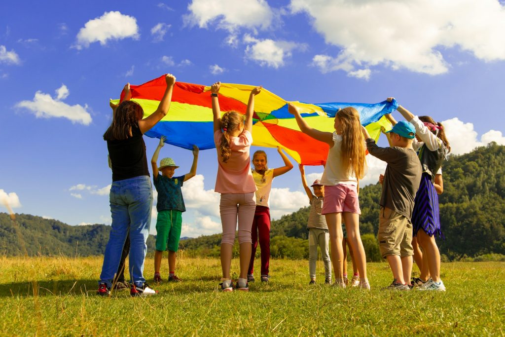 Eine Gruppe von Kindern spielt im Freien mit einem bunten Fallschirm auf einer Wiese. Der Himmel ist blau mit einigen Wolken, und im Hintergrund sind sanfte Hügel zu sehen. Die Kinder zeigen Freude und Teamarbeit, während sie den Fallschirm hochhalten.