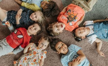 Gruppe von acht fröhlichen Kindern, die auf einem Teppich liegen und in die Kamera lächeln. Sie tragen bunte Kleidung und zeigen eine entspannte, spielerische Atmosphäre.