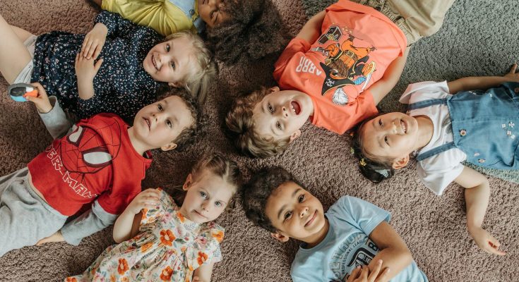 Gruppe von acht fröhlichen Kindern, die auf einem Teppich liegen und in die Kamera lächeln. Sie tragen bunte Kleidung und zeigen eine entspannte, spielerische Atmosphäre.