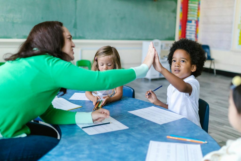 Eine Lehrerin gibt einem Schüler im Klassenzimmer ein High-Five, während zwei weitere Kinder am Tisch arbeiten. Die Szene zeigt eine positive Lernumgebung mit Interaktion und Zusammenarbeit.