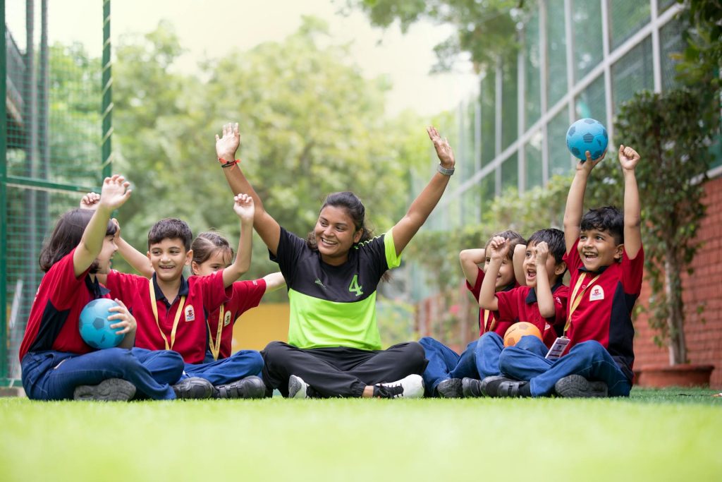 Eine Gruppe von Kindern in roten T-Shirts sitzt lachend auf einer Wiese, während eine Trainerin in einem grünen Sportoutfit begeistert die Arme hebt. Die Kinder halten verschiedene Bälle in den Händen und zeigen Freude und Begeisterung. Im Hintergrund sind Bäume und ein Sportplatz sichtbar.