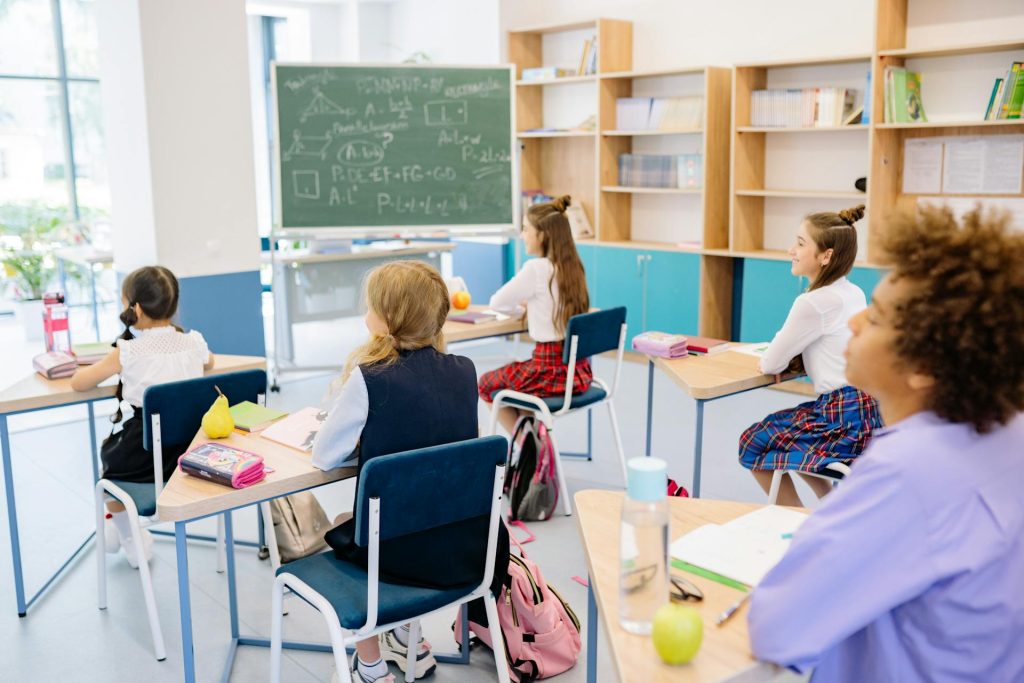Schülerinnen und Schüler sitzen in einem modernen Klassenzimmer und hören dem Unterricht an. Auf dem Tisch liegen Bücher, Schreibmaterial und Snacks. Im Hintergrund ist eine Tafel mit mathematischen Formeln zu sehen.
