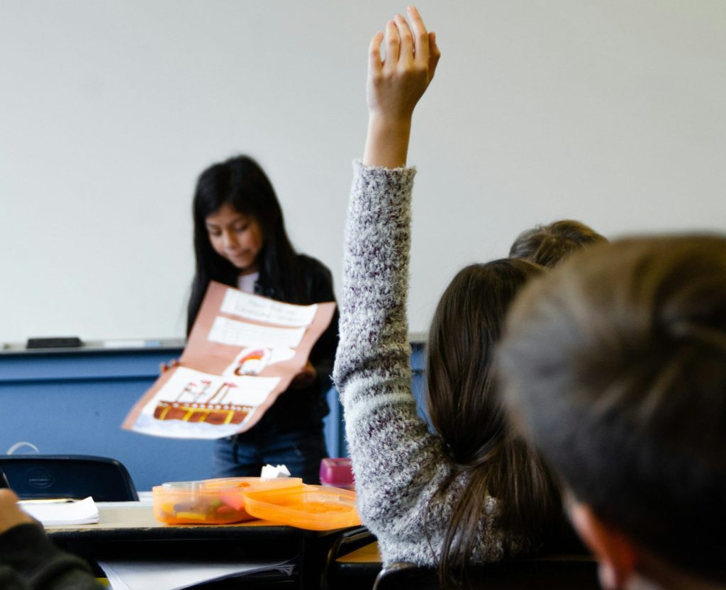 Schüler im Klassenzimmer heben die Hand, während eine Mitschülerin ein Projekt präsentiert. Auf dem Tisch sind Materialien sichtbar, die zur Präsentation genutzt werden.