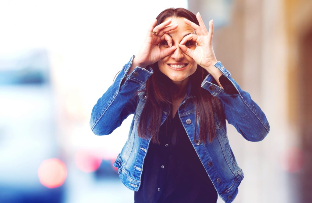 Junge Frau in einer Jeansjacke und schwarzem Oberteil, die mit ihren Händen eine lustige Geste macht, während sie lächelt. Unscharfer urbaner Hintergrund mit verschwommenen Lichtern.