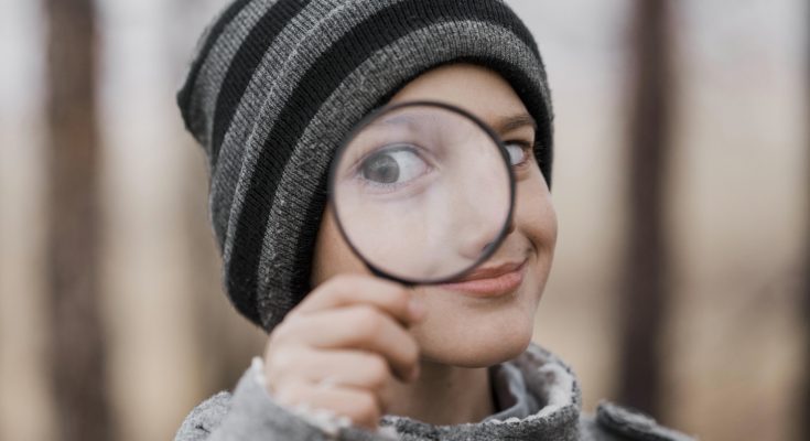 Ein Junge mit einem gestreiften Mütze hält eine Lupe vor sein Auge und schaut neugierig. Der Hintergrund ist unscharf und zeigt eine natürliche Umgebung.