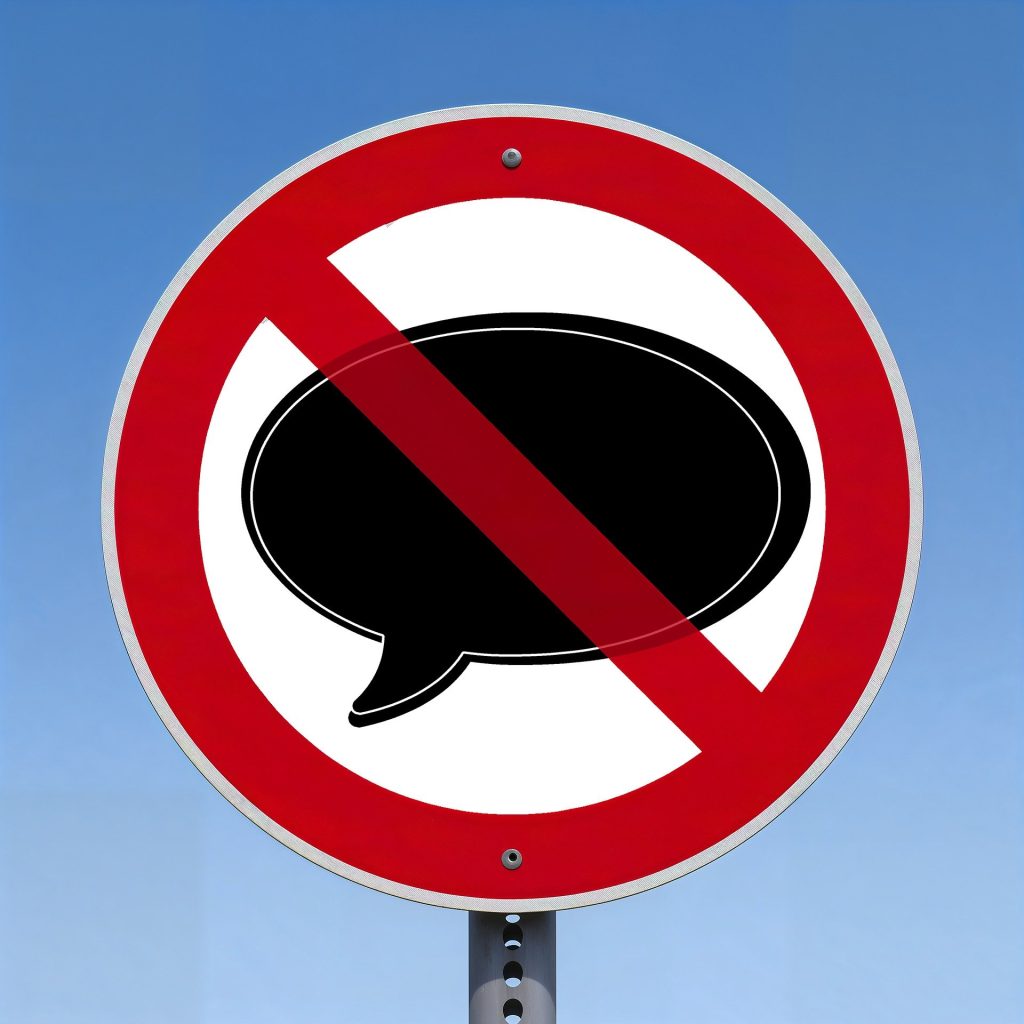 Verkehrsschild mit rotem Rand und durchgestrichenem Sprechblasen-Symbol, das das Verbot von Kommunikation oder Lautäußerungen signalisiert, vor klarem blauen Himmel.