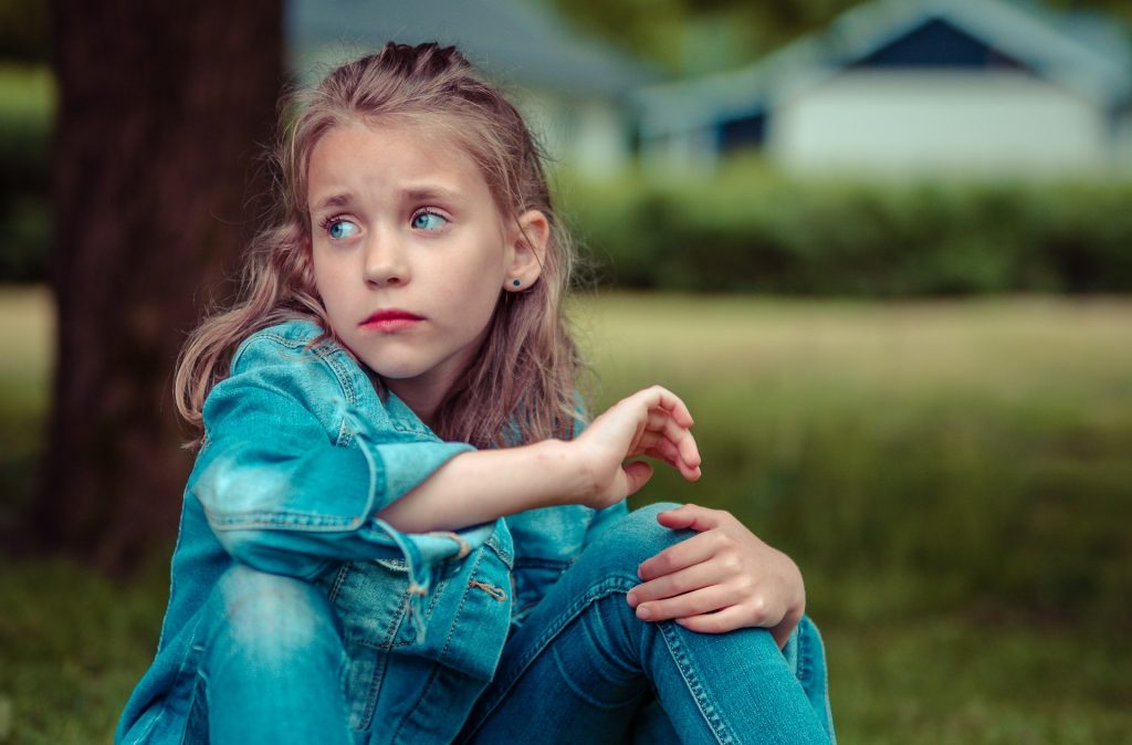 Ein nachdenkliches Mädchen sitzt auf dem Gras und trägt eine blaue Jeansjacke sowie passende Jeans. Ihr Blick ist nachdenklich und sie hält ihre Beine umschlungen. Im Hintergrund sind unscharfe Bäume und Gebäude zu sehen, was eine ruhige, natürliche Umgebung schafft.