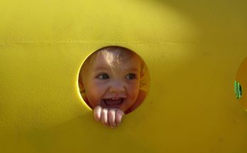Ein fröhliches Kleinkind schaut durch ein rundes Loch in einem gelben Spielgerät, mit einem breiten Lächeln und strahlenden Augen. Das Bild vermittelt Freude und Unbeschwertheit beim Spielen im Freien.