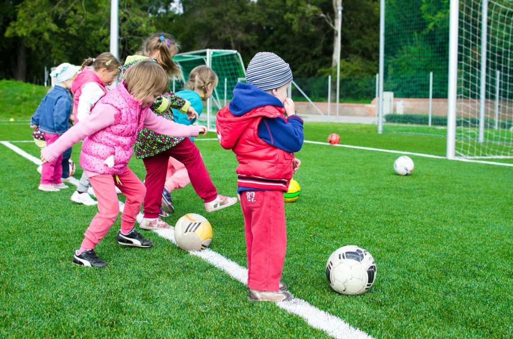 Gruppe von Kindern, die auf einem Fußballfeld spielen. Die Kinder tragen bunte Sportkleidung und stehen in einer Reihe, bereit, den Ball zu kicken. Im Hintergrund sind Tore und weitere Fußbälle sichtbar.