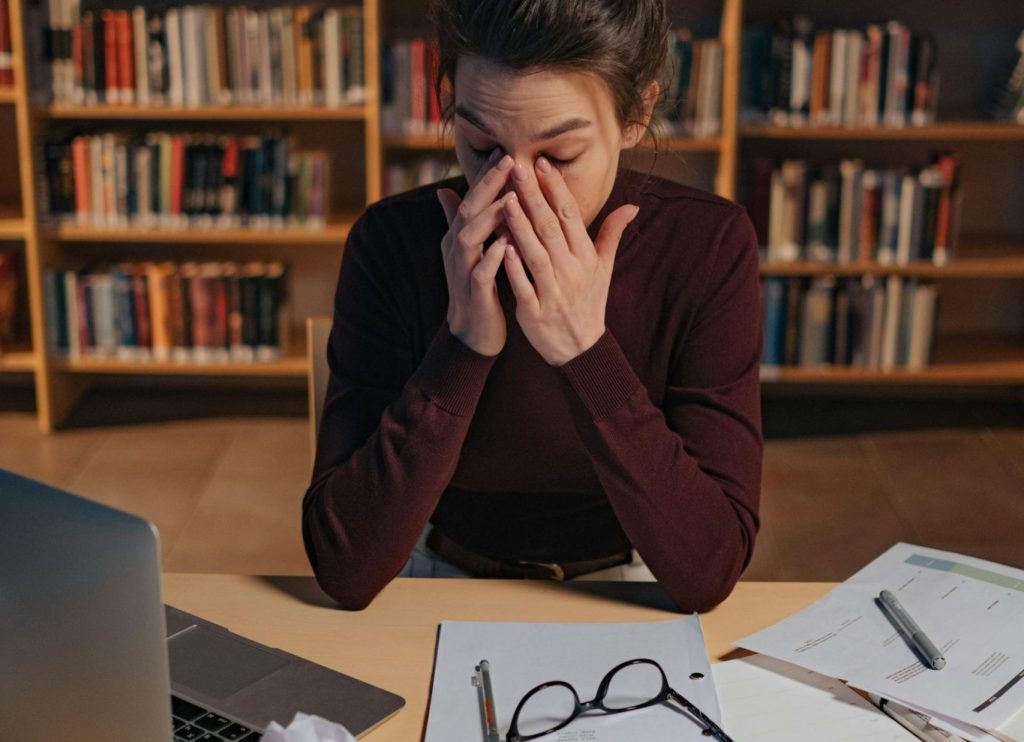Eine gestresste Frau sitzt an einem Schreibtisch in einer Bibliothek, hält sich das Gesicht mit beiden Händen und sieht frustriert aus. Um sie herum liegen zerknüllte Papiere, Notizen und ein Laptop, was auf eine intensive Arbeitsatmosphäre hindeutet.