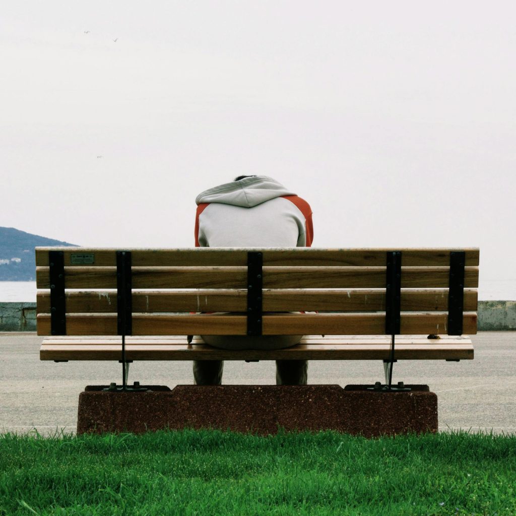 Eine Person sitzt auf einer Holzbank mit Blick auf das Wasser, umgeben von einer grünen Wiese. Die Stimmung ist ruhig und nachdenklich, ideal für Entspannung oder Reflexion.