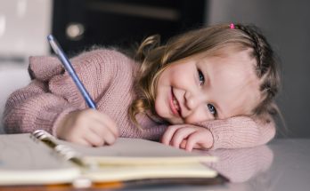 Ein lächelndes kleines Mädchen sitzt an einem Tisch, während sie mit einem blauen Stift in ein Notizbuch schreibt. Sie trägt einen rosa Pullover und hat ihre Arme auf den Tisch gelegt. Die Umgebung ist hell und modern, was eine freundliche und kreative Atmosphäre vermittelt.