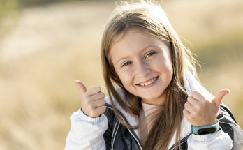 Ein lächelndes Mädchen mit langen, blonden Haaren gibt zwei Daumen nach oben. Sie trägt eine weiße Jacke und einen Fitness-Tracker am Handgelenk. Im Hintergrund sind unscharfe, goldene Gräser zu sehen, die eine fröhliche und positive Stimmung vermitteln.