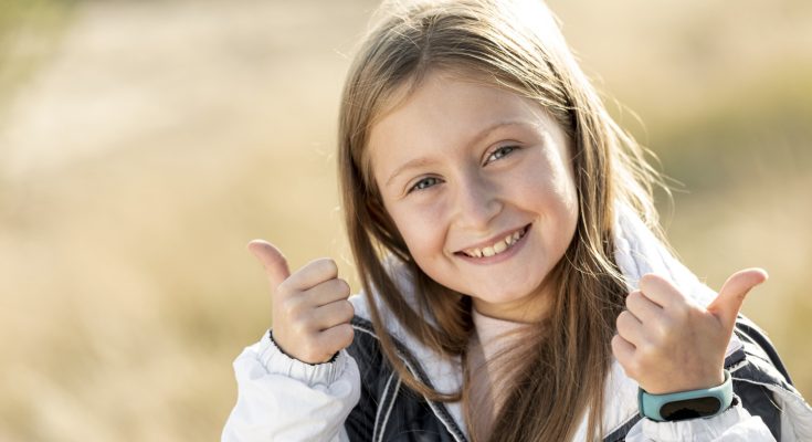 Ein lächelndes Mädchen mit langen, blonden Haaren gibt zwei Daumen nach oben. Sie trägt eine weiße Jacke und einen Fitness-Tracker am Handgelenk. Im Hintergrund sind unscharfe, goldene Gräser zu sehen, die eine fröhliche und positive Stimmung vermitteln.