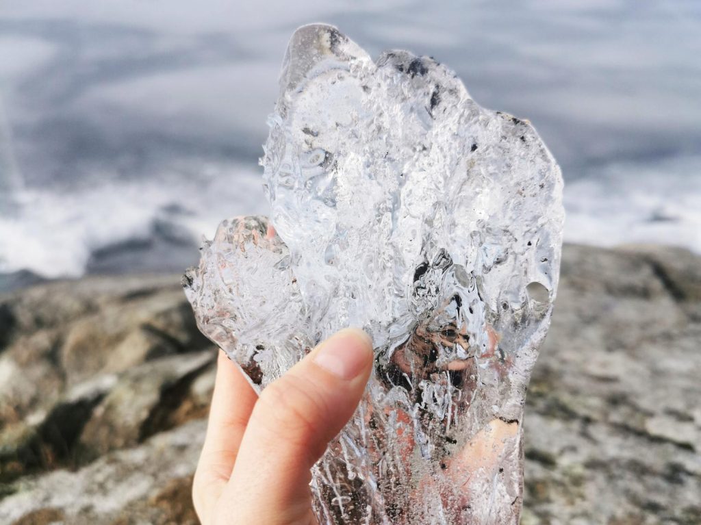 Eine Hand hält ein großes, transparentes Eisstück vor einer ruhigen Wasseroberfläche und steiniger Küste. Das Eis zeigt interessante Strukturen und reflektiert das Licht. Ideal für Themen wie Natur, Winter oder Eiskunst.