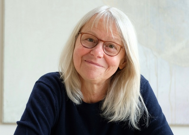 Porträt einer lächelnden Frau mit langen, grauen Haaren und Brille, die in einem hellen, modernen Raum sitzt. Sie trägt einen dunklen Pullover und strahlt Freundlichkeit aus. Im Hintergrund sind abstrakte Kunstwerke sichtbar.