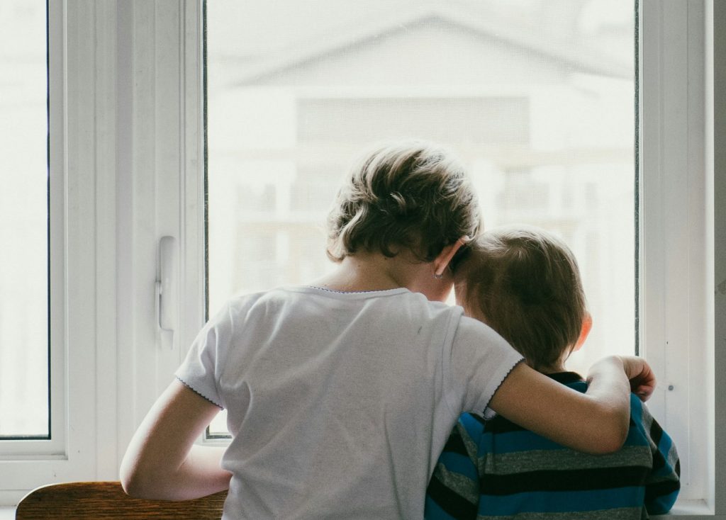 Zwei Kinder stehen eng umschlungen vor einem Fenster und schauen nach draußen. Die Szene vermittelt ein Gefühl von Nähe und Freundschaft in einem hellen, ruhigen Raum.