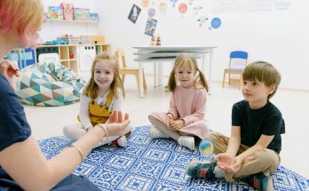 In einem hellen und einladenden Klassenzimmer sitzen drei Kinder auf einem blauen Teppich. Sie schauen gespannt auf eine Person, die ihnen etwas zeigt. Im Hintergrund sind Spielzeug und Lernmaterialien sichtbar, die eine kreative Lernumgebung schaffen.
