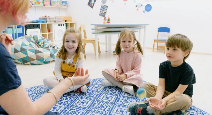 In einem hellen und einladenden Klassenzimmer sitzen drei Kinder auf einem blauen Teppich. Sie schauen gespannt auf eine Person, die ihnen etwas zeigt. Im Hintergrund sind Spielzeug und Lernmaterialien sichtbar, die eine kreative Lernumgebung schaffen.