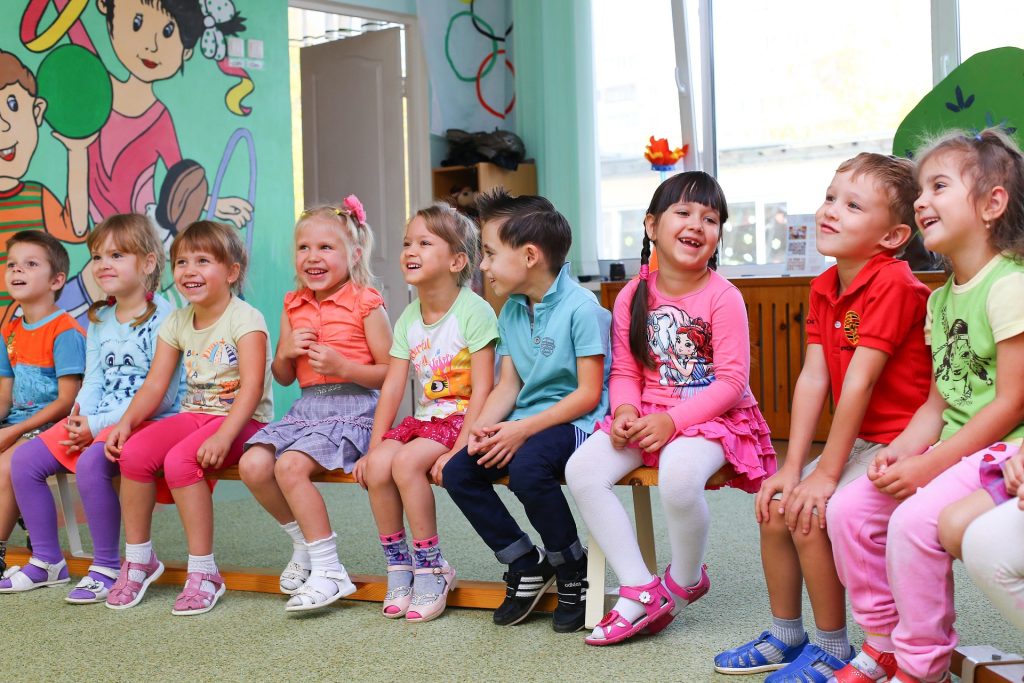 Eine Gruppe fröhlicher Kinder sitzt auf einer Bank in einem bunten Raum. Sie lachen und schauen einander an, während sie in verschiedenen bunten T-Shirts und Hosen gekleidet sind. Im Hintergrund sind kindgerechte Wandmalereien zu sehen.