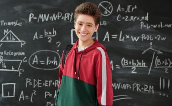 Junger Schüler lächelt vor einer Tafel mit mathematischen Formeln und Geometriezeichnungen, während er einen roten und grünen Hoodie trägt.
