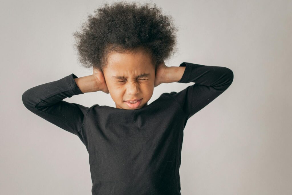 Ein Kind mit lockigem Haar hält sich die Ohren zu und zeigt einen Ausdruck von Unbehagen. Der Hintergrund ist neutral und hell, was die Emotionen des Kindes hervorhebt.