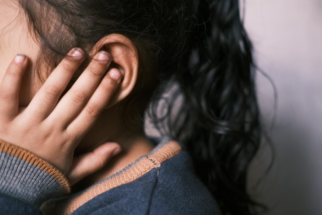 Ein Kind hält sich die Hand ans Ohr und zeigt eine nachdenkliche oder besorgte Miene. Die Aufnahme fokussiert auf die Hand und das Ohr, während das Gesicht teilweise verdeckt ist. Die Kleidung des Kindes ist in sanften Farben gehalten.