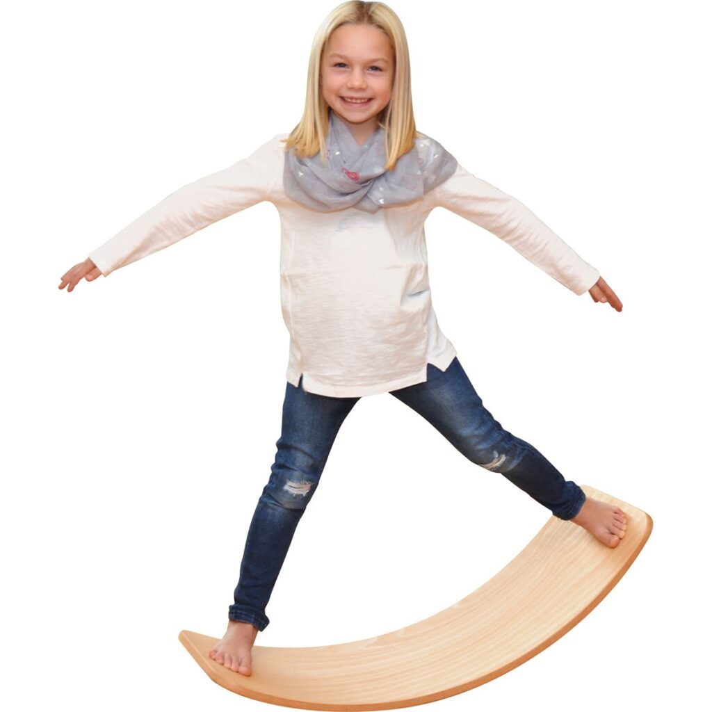 Ein kleines Mädchen balanciert fröhlich auf einem Holzspielgerät. Sie trägt ein weißes Oberteil und eine graue Schal, während sie mit beiden Armen für Gleichgewicht sorgt. Das Bild vermittelt Spaß und Bewegungsfreude für Kinder.