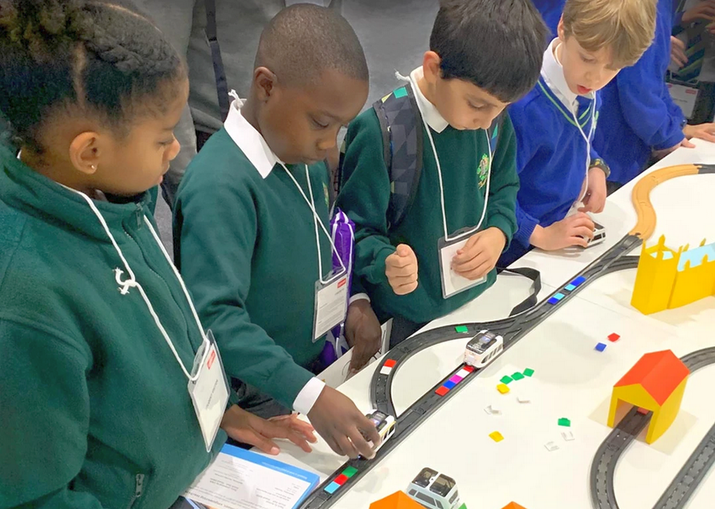 Gruppe von Kindern, die an einem Tisch mit einer Modellbahn spielen und verschiedene Fahrzeuge steuern. Die Kinder sind in Schulkleidung und konzentrieren sich auf das Spiel, während sie mit bunten Bausteinen und einer Spielbahn interagieren.