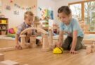 Zwei Kinder spielen in einem hellen Klassenraum mit Holzbausteinen. Ein Junge untersucht eine Baukonstruktion, während ein Mädchen einen programmierbaren Roboter in Form einer Biene steuert. Im Hintergrund sind farbenfrohe Kunstwerke und Spielmaterialien sichtbar, die eine kreative Lernumgebung schaffen.
